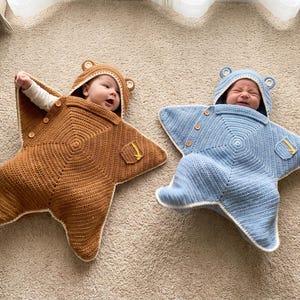 May include: Two infants wearing star-shaped crochet swaddles with hoods featuring small ears. One swaddle is brown and the other is light blue, both with white trim, wooden buttons, and a small pocket on the front. The crochet pattern radiates from the center.