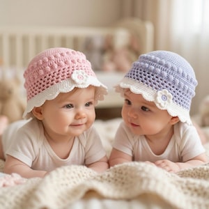 May include: Two crochet baby hats, one in light pink and one in light purple, both featuring a white scalloped brim and a small white flower accent. The hats have a textured bobble stitch pattern on the crown and a grid-like weave on the sides.