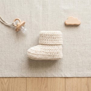 Pu&ograve; includere: Un stivaletto da bambino all'uncinetto color crema con risvolto &egrave; appoggiato su una superficie di lino neutra. Sono presenti anche un ciuccio in legno e un giocattolo di legno a forma di nuvola. Lo stivaletto &egrave; probabilmente fatto a mano.