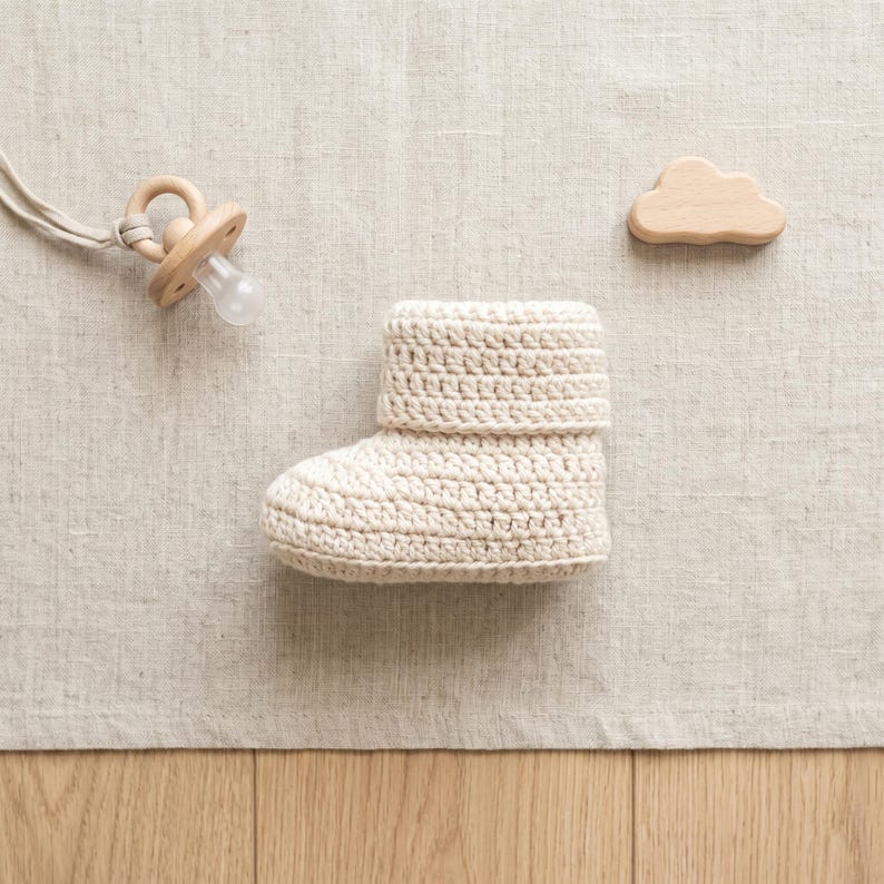 Pu&ograve; includere: Un stivaletto da bambino all'uncinetto color crema con risvolto &egrave; appoggiato su una superficie di lino neutra. Sono presenti anche un ciuccio in legno e un giocattolo di legno a forma di nuvola. Lo stivaletto &egrave; probabilmente fatto a mano.