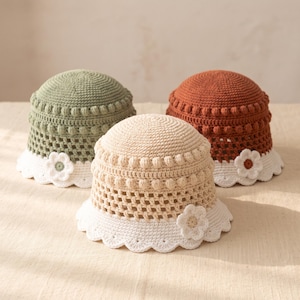 May include: Three crochet bucket hats in sage green, beige, and terracotta orange. Each hat features a textured bobble stitch band, a lattice-patterned middle section, and a white scalloped brim adorned with a single crocheted flower applique.
