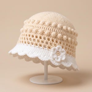 May include: A cream-colored crochet bucket hat featuring a textured bobble stitch pattern around the crown, a lattice-style openwork middle section, and a white scalloped brim adorned with a single crocheted flower accent on the side.