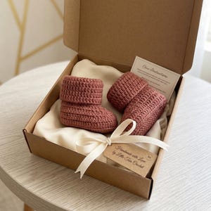 Pu&ograve; includere: Un paio di stivaletti da neonato all'uncinetto rosa antico fatti a mano, presentati in una scatola regalo di cartone. Gli stivaletti sono appoggiati su un tessuto color crema e legati con un nastro color crema. Una piccola etichetta recita "Handmade with Love by Little Toes Crochet".