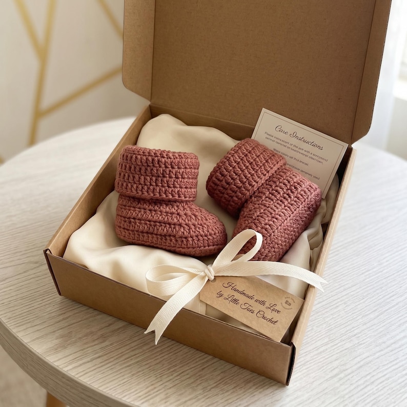Pu&ograve; includere: Un paio di stivaletti da neonato all'uncinetto rosa antico fatti a mano, presentati in una scatola regalo di cartone. Gli stivaletti sono appoggiati su un tessuto color crema e legati con un nastro color crema. Una piccola etichetta recita "Handmade with Love by Little Toes Crochet".