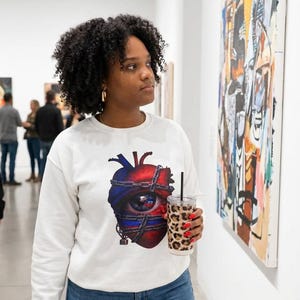 May include: White sweatshirt with a graphic of a heart and eye design. The person is holding a leopard print tumbler with a black straw. The person is wearing blue jeans and is in an art gallery.