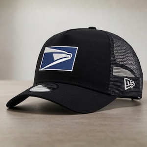 May include: Black trucker hat featuring the USPS logo embroidered on the front. The logo is a white eagle within a blue rectangle. The hat has a curved brim and a mesh back panel. A New Era logo is on the side.