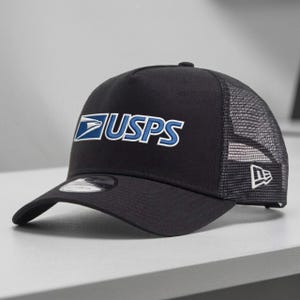 May include: Black trucker hat with the USPS logo in white and blue. The hat features a mesh back and a curved brim. The New Era logo is on the side.