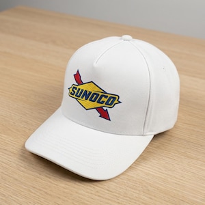 May include: A white baseball cap featuring the Sunoco logo in yellow, blue, and red. The logo is embroidered on the front of the cap, which has a curved brim and a structured crown. The cap is made of a durable fabric.