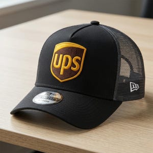 May include: Black trucker hat with a mesh back and a curved brim. The front features a gold and brown UPS logo patch. A New Era sticker is on the brim. The hat has a snapback closure.
