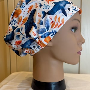 May include: A white scrub cap with a marine life pattern. The design features blue whales, orange fish, and blue and orange foliage. The cap is made of a soft fabric and is designed for comfort.