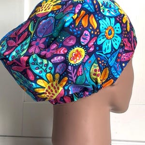 May include: A patterned bouffant surgical scrub cap. The cap features a vibrant floral design with blue, yellow, pink, and purple flowers and leaves on a dark blue background. The cap is designed to cover the hair.