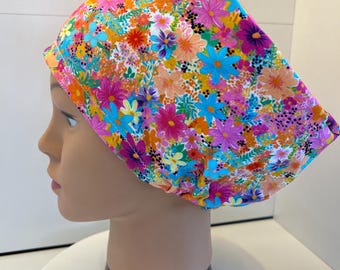 May include: A scrub cap featuring a vibrant, multi-colored floral pattern on a white background. The fabric displays a dense arrangement of small flowers in shades of bright pink, orange, turquoise, yellow, and purple, with green foliage accents.