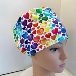 Può includere: Una cuffia chirurgica con sfondo bianco e un motivo ripetuto di cuori in stile acquerello in vari colori, tra cui rosso, arancione, giallo, verde, blu e viola. La cuffia in tessuto ha una forma arrotondata e un orlo cucito lungo il bordo.