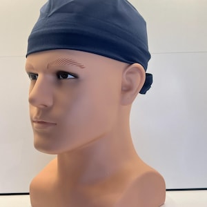 May include: A solid navy blue scrub cap featuring a fitted crown and a wide, smooth headband section that wraps around the forehead. The fabric is plain and matte, with a small tie closure visible at the back of the head on the mannequin.