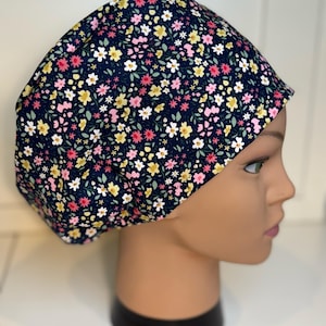 May include: A navy blue scrub cap adorned with a floral design. The cap displays a repeating pattern of small pink, yellow, and white flowers with green leaves. The cap is designed to cover the head and hair.