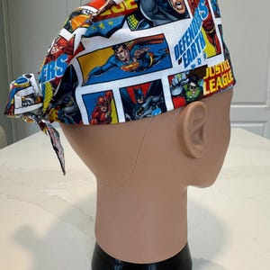 Puede incluir: Un gorro quirúrgico colorido con paneles de cómic con superhéroes como Batman, Superman y Flash. El gorro tiene un cierre de corbata y las palabras "DEFENDERS OF EARTH" y "JUSTICE LEAGUE".