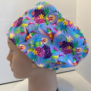 May include: A scrub cap featuring a light purple background with a vibrant pattern of colorful hummingbirds, pink dahlias, yellow flowers, and calla lilies. The fabric design includes green foliage and tropical leaves distributed across the entire surface.