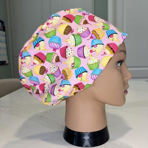 May include: A scrub cap featuring a light pink background with a repeating pattern of colourful cupcakes. Each cupcake has various coloured frosting, including blue, yellow, purple, and pink, with some topped with a small red cherry illustration.