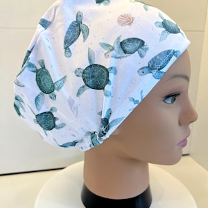 Könnte beinhalten: Eine weiße OP-Haube mit einem wiederkehrenden Muster aus grünen Meeresschildkröten und kleinen rosa Muscheln. Die Kopfbedeckung aus Stoff hat eine abgerundete Form mit gerafftem Hinterkopf und sichtbaren Nähten am unteren Rand.