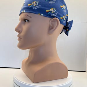May include: A blue scrub cap featuring a repeating pattern of the Millennium Falcon spaceship with a rainbow trail and the text Millennium Falcon printed in white. The cap is tied at the back with fabric ends and has a fitted, rounded crown shape.