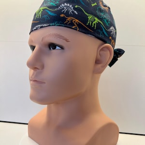 May include: A dark navy blue scrub cap featuring a repeating pattern of colourful dinosaur skeletons in shades of lime green, orange, and light blue. The cap is fitted with a tie-back closure at the rear and covers the top of the head.