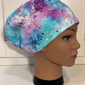 Può includere: Un cappello chirurgico colorato con un design ad acquerello. Il cappello presenta una miscela di tonalità blu, viola e bianche, accentuata da dettagli di stelle e macchie dorate. Il cappello è esposto su una testa di manichino.