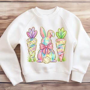 May include: White sweatshirt featuring a colorful Easter bunny design with a pink bow and two carrots. The bunny and carrots have a leopard print pattern. The design is set against a wooden background.