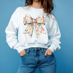 Peut inclure: Sweat-shirt blanc orné d'un grand nœud orné d'œufs de Pâques colorés. Le nœud a un fond crème avec des œufs pastel roses, jaunes, bleus et verts. Le modèle porte un jean bleu.