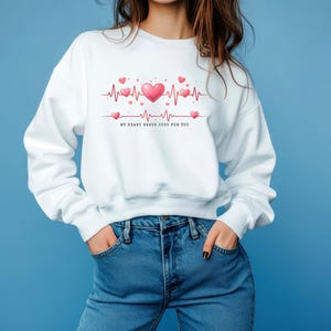 以下が含まれることがあります： ハートをテーマにしたデザインの白いスウェットシャツ。グラフィックは、ピンクのハートと「MY HEART BEATS JUST FOR YOU」のテキストが入った心拍ラインが特徴です。モデルはブルージーンズを着用しています。