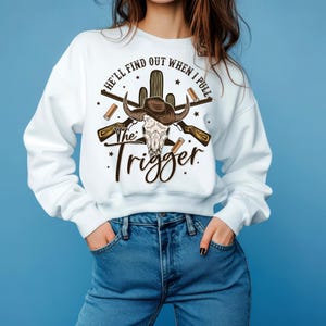 May include: A white long-sleeve sweatshirt featuring a graphic print of a bull skull wearing a cowboy hat, crossed rifles, a cactus, and shotgun shells. The text reads, "HE'LL FIND OUT WHEN I PULL The Trigger" in black and brown lettering.