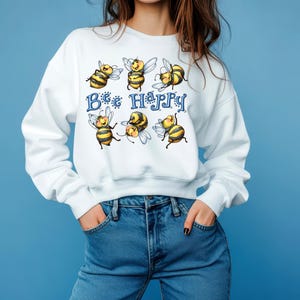 Może przedstawiać: Biała bluza z nadrukiem "Bee Happy". Grafika przedstawia kilka kreskówkowych pszczół w kolorze żółtym i czarnym, z niebieskimi literami. Osoba ma na sobie niebieskie dżinsy.