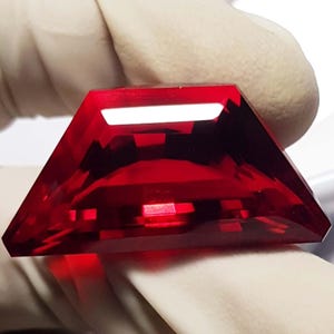 May include: A faceted, trapezoid-shaped gemstone with a deep red hue. The gem is held by a gloved hand, showcasing its polished surfaces and internal reflections. The cut of the stone allows light to pass through, creating a brilliant display.