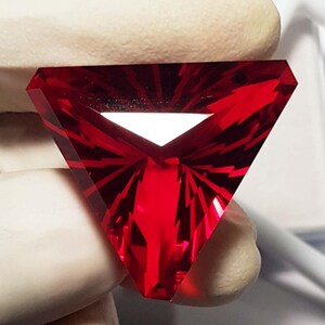 May include: A faceted, triangular gemstone with a deep red hue. The gem features a geometric design with sharp angles and a central, white, triangular void. The light reflects off the facets, creating a brilliant display.
