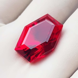 May include: A faceted, rectangular-cut, deep red gemstone. The gem has a complex, multi-faceted design that reflects light, creating a brilliant shine. The gemstone is set against a white background.