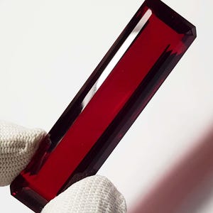 May include: A rectangular, faceted, deep red gemstone is held by a gloved hand. The gem has a polished, transparent appearance, with sharp edges and a rich, saturated color. The gem's shape is elongated, with a flat top and bottom.