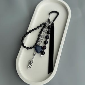 Op de afbeelding: Een verzameling zwarte kralenjuwelen en een zilveren ketting met een blauwe hartbedel en een kleine berenbedel. De sieraden zijn bevestigd aan een zwarte karabijnhaak en een zwart lint, alles op een witte ovale tray.