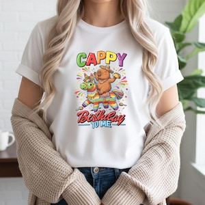 Op de afbeelding: Wit T-shirt met een kleurrijke afbeelding van een beer die op een piñata rijdt. De tekst op het shirt luidt "Cappy Birthday To Me" in een speels lettertype. Het ontwerp bevat levendige kleuren en confetti, wat een feestelijke uitstraling creëert.