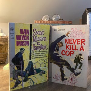 May include: Two paperback books with yellow covers. The book on the left is titled "Secret Mission to Bangkok" and features a man in a blue suit standing over a man lying on a bed. The book on the right is titled "Never Kill a Cop" and features a man in a blue suit jumping over another man in a brown suit. Both books are part of the "Van Wyck Mason" series.