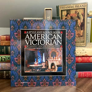May include: A hardcover book titled "American Victorian" with a blue and pink patterned cover. The book's cover features an interior scene and the text "A Style and Source Book". Other books are visible in the background, including one titled "Treasure Island".