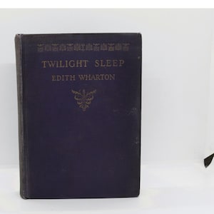 May include: A dark purple hardcover book titled Twilight Sleep by Edith Wharton. The front cover features gold-colored text and a decorative floral-style emblem centered below the author's name, with a repeating pattern along the top edge.