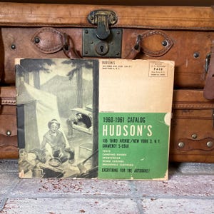 May include: A vintage Hudson's catalog from 1960-1961, featuring a green and white cover with illustrations of camping scenes. The catalog lists tents, camping goods, sportswear, work clothes, and industrial clothing. The catalog is placed in front of a brown leather suitcase.