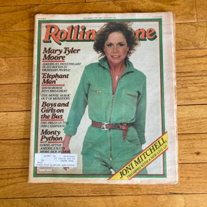 May include: A vintage Rolling Stone magazine cover featuring Mary Tyler Moore in a green jumpsuit. The magazine's masthead is red and green. Other text includes "Elephant Man" and "Joni Mitchell Triumphs with a New Live LP."