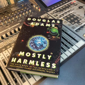 May include: A black and green book with the title "Mostly Harmless" by Douglas Adams. The book is the fifth in the Hitchhiker's Guide to the Galaxy series.