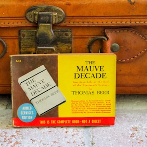 May include: A vintage book titled "The Mauve Decade" by Thomas Beer, an Armed Services Edition. The book has a yellow and brown cover with text and is set against a brown leather suitcase.