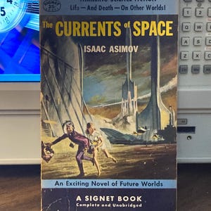 May include: Vintage paperback book titled "The Currents of Space" by Isaac Asimov. The cover features a futuristic cityscape with running figures. The book is a Signet Book, complete and unabridged, with the text "Thrilling Science Fiction" and "An Exciting Novel of Future Worlds".