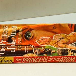 May include: A vintage paperback book titled "The Princess of the Atom" by Ray Cummings. The cover features a woman's eye and a portion of her face, along with a futuristic-looking device.