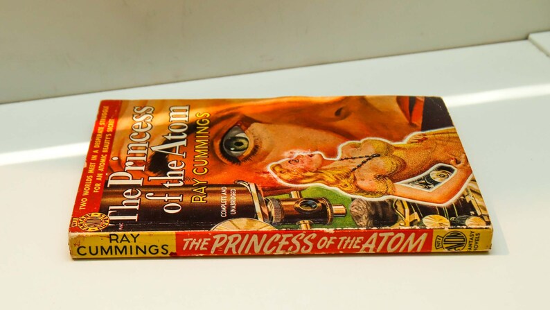 May include: A vintage paperback book titled "The Princess of the Atom" by Ray Cummings. The cover features a woman's eye and a portion of her face, along with a futuristic-looking device.