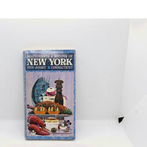 May include: A rectangular cookbook titled Restaurants & Recipes of New York, New Jersey, Connecticut. The cover features a blue and purple illustration of a fish, a pepper mill, a roasted bird, and lobster claws on a serving platter.