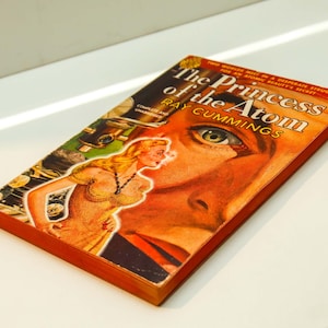 May include: A vintage paperback book titled "The Princess of the Atom" by Ray Cummings. The cover features a woman in a yellow dress and a man's face with a green eye.