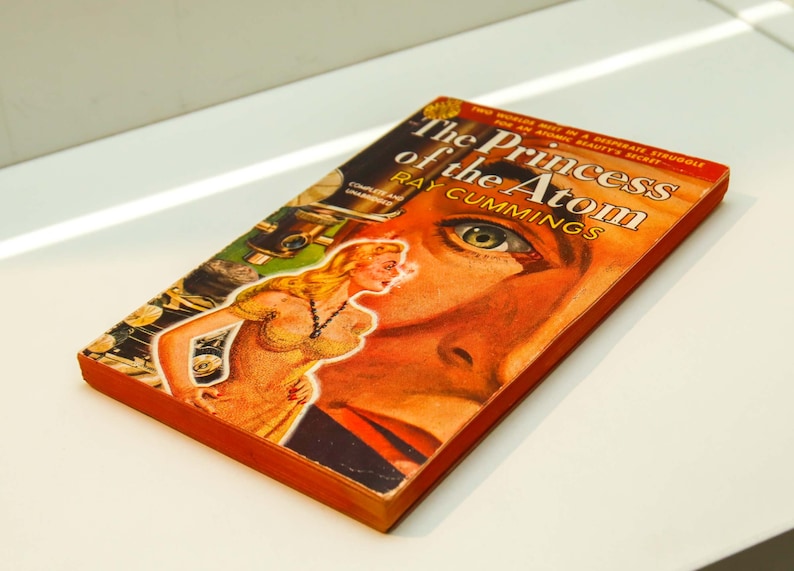 May include: A vintage paperback book titled "The Princess of the Atom" by Ray Cummings. The cover features a woman in a yellow dress and a man's face with a green eye.